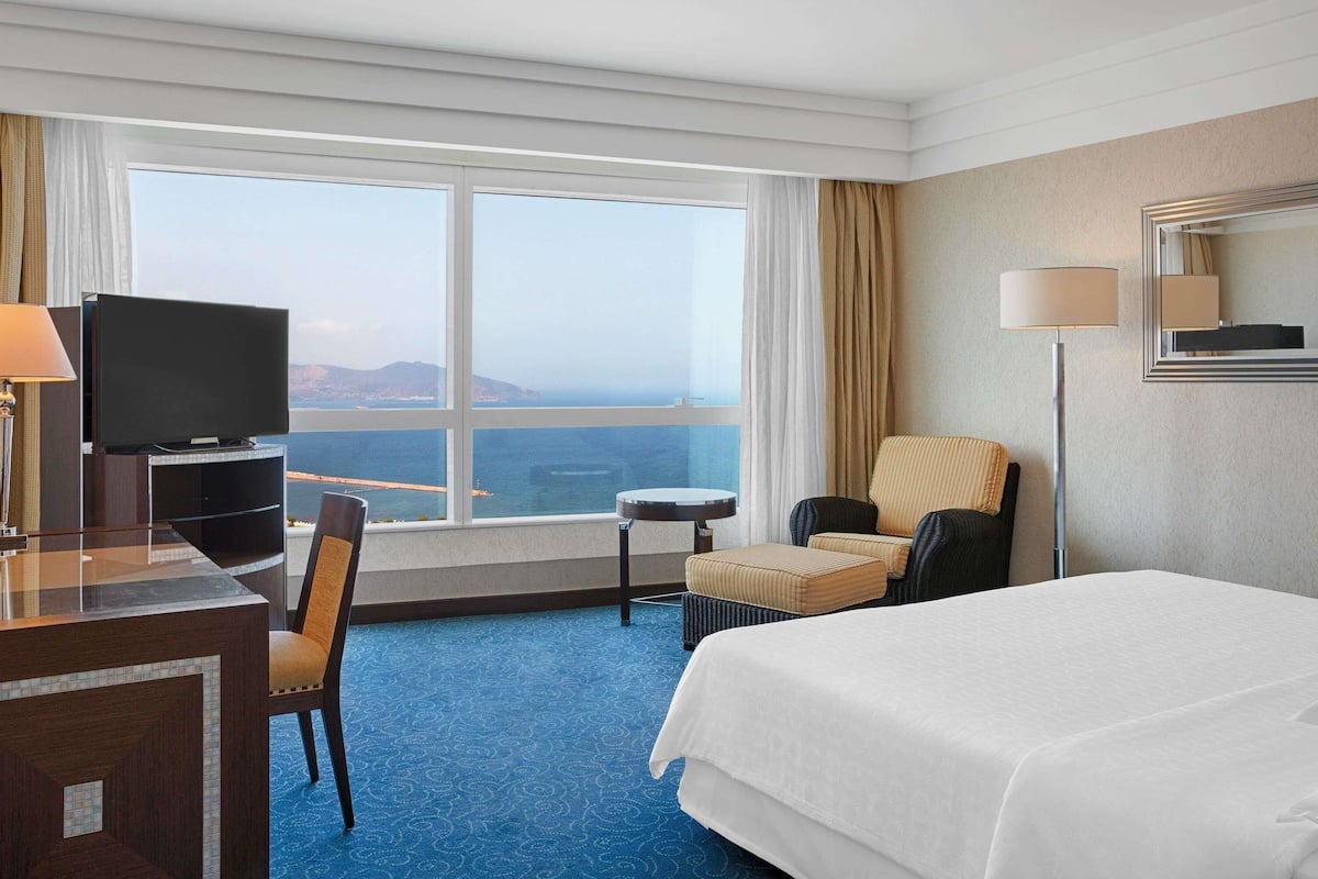 A hotel room with a bed, a desk with a television, a chair, a small table, and a view of the sea.