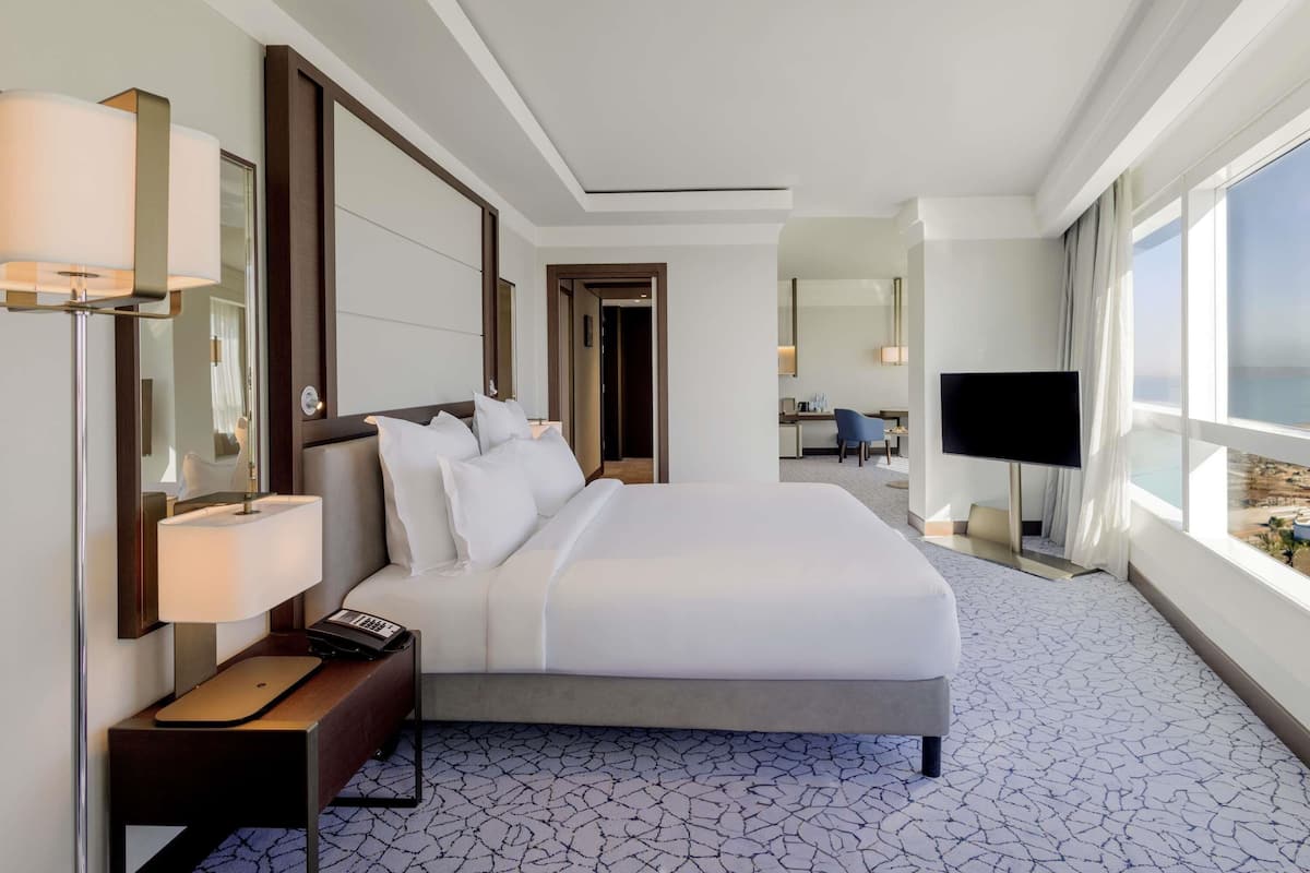 A hotel room with a large bed, bedside lamp, desk, chair, TV, and a view of the sea.