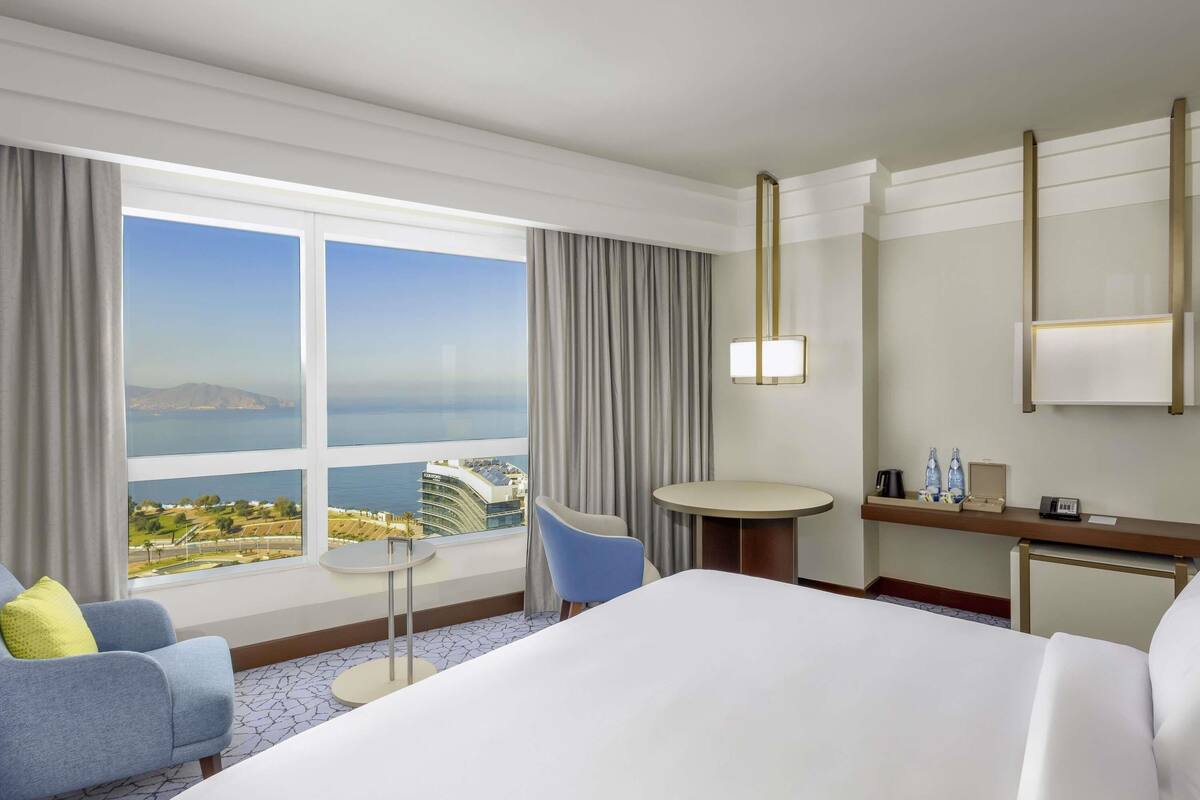 A hotel room with a large bed, a desk, a chair, and a view of a cityscape through large windows.