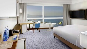 A hotel room with a large bed, a desk with a chair, a TV mounted on the wall, and a view of the sea through a large window. - Hotel Oran Bay Managed By Accor (Oran)