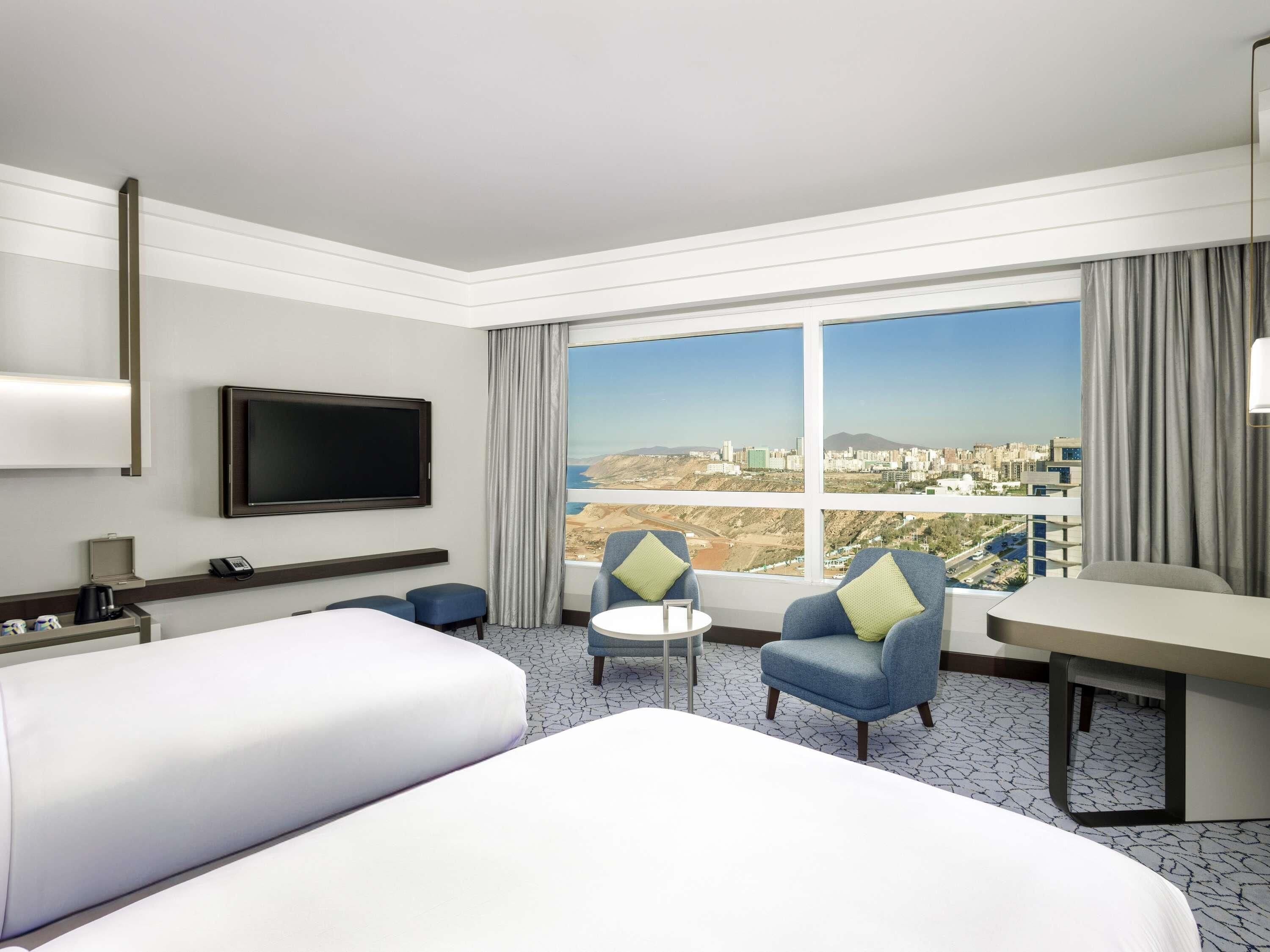 Ein Hotelzimmer mit Bett, zwei Sesseln, einem kleinen Tisch, einem Fernseher und einem großen Fenster mit Blick auf die Stadt.
