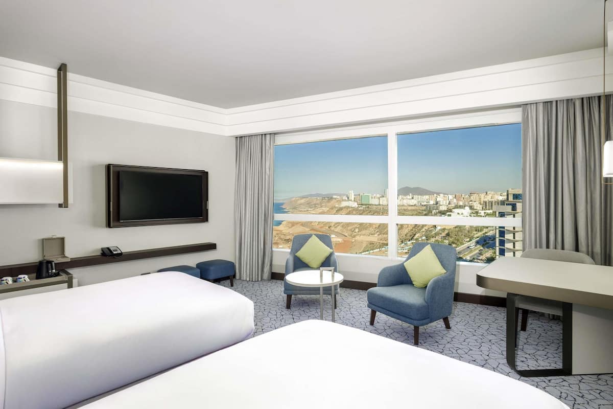 A hotel room with a bed, two armchairs, a small table, a TV, and a large window with a city view.