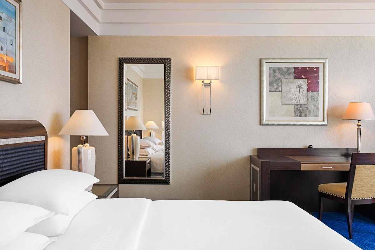 A hotel room with a bed, bedside lamps, a desk, a chair, a mirror, and a framed picture on the wall.