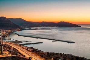 A coastal cityscape at sunset with a prominent road leading to a waterfront area, featuring a large body of water, a pier, and distant mountains. - Hotel Oran Bay Managed By Accor (Oran)