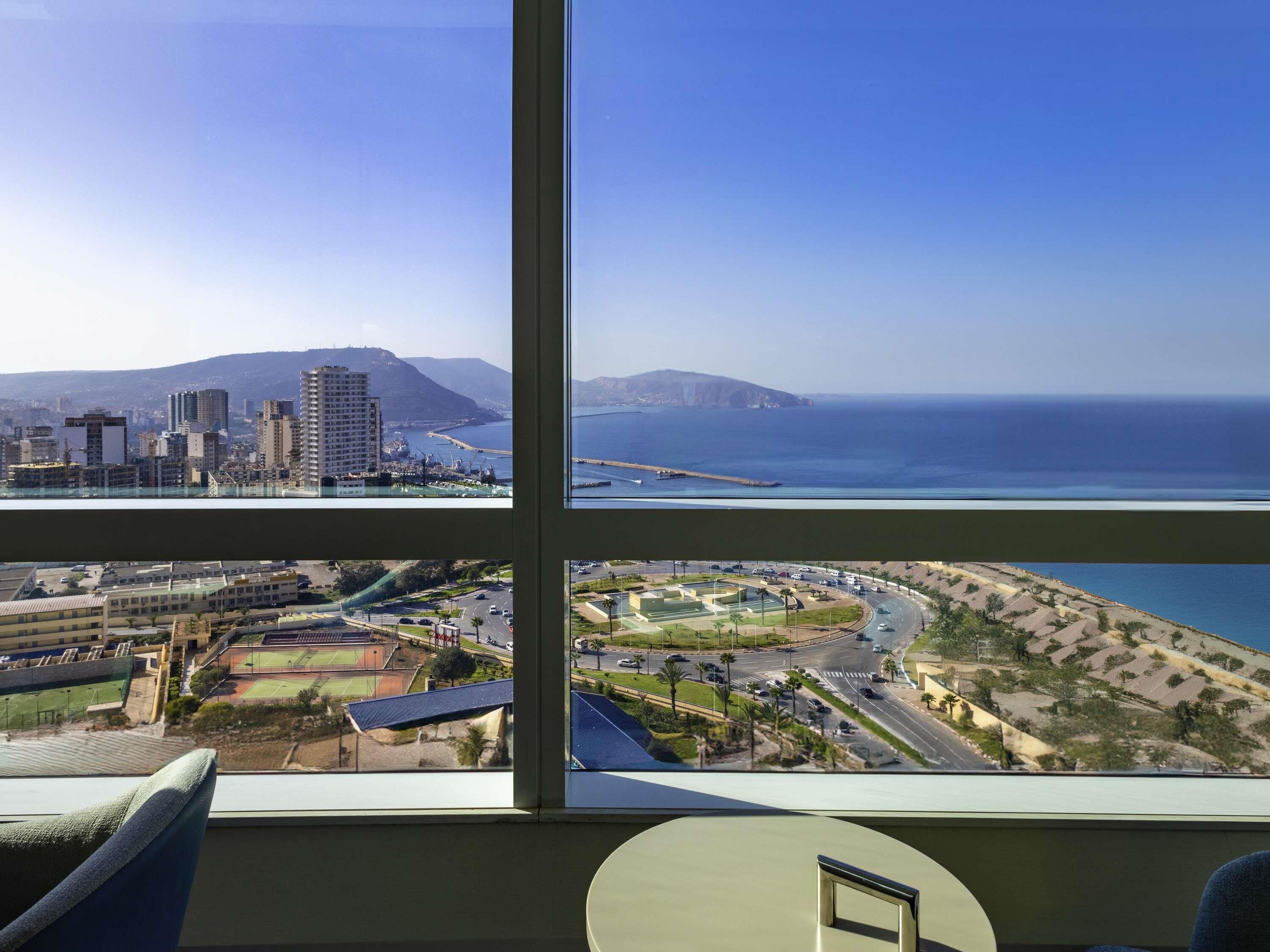 Habitación de hotel en un edificio alto con un ventanal grande que ofrece una vista panorámica de una ciudad costera, incluyendo un complejo deportivo y una carretera con vehículos.