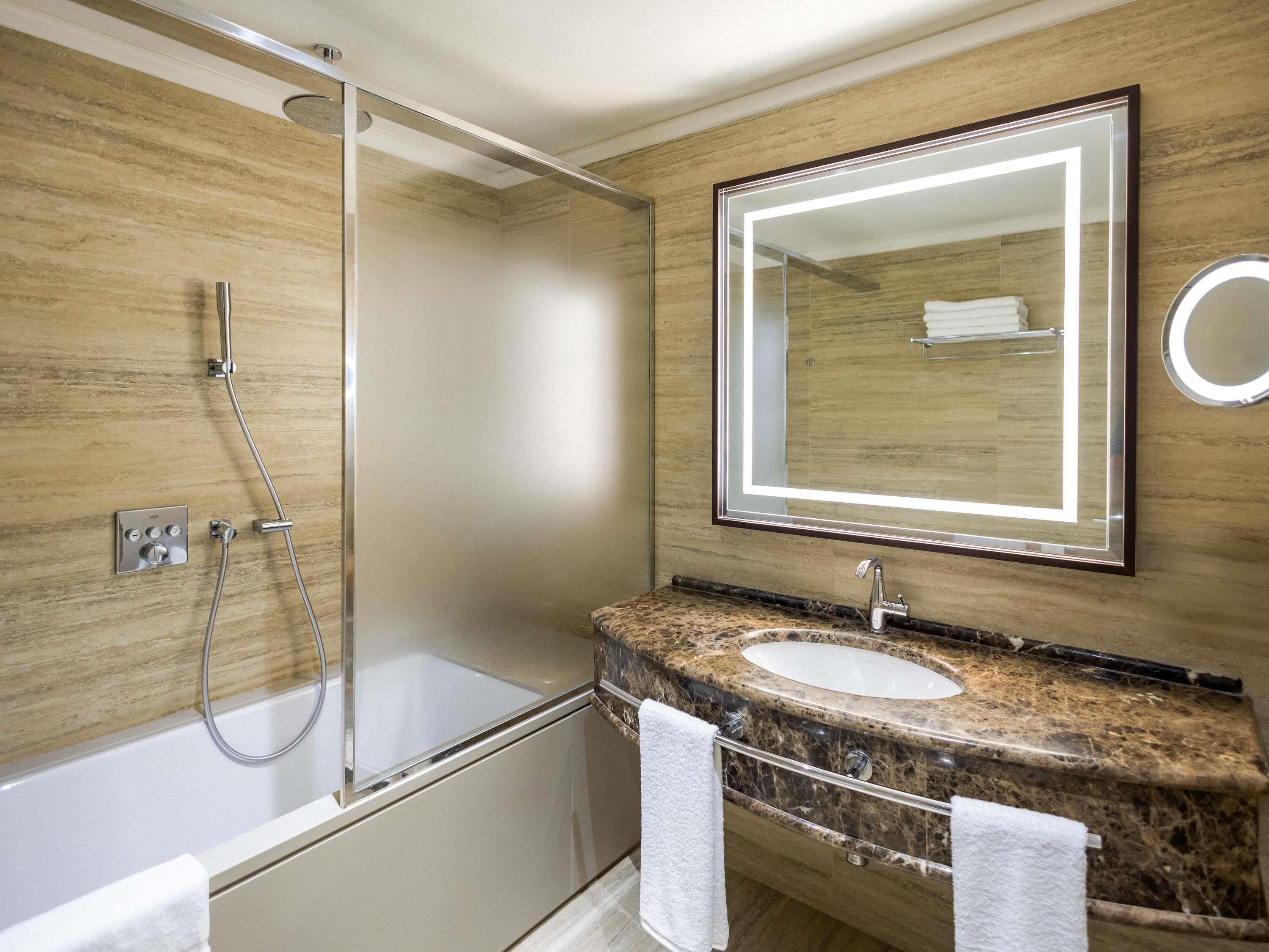 A modern bathroom with a glass-enclosed shower, a marble sink, and a large mirror with built-in lighting.