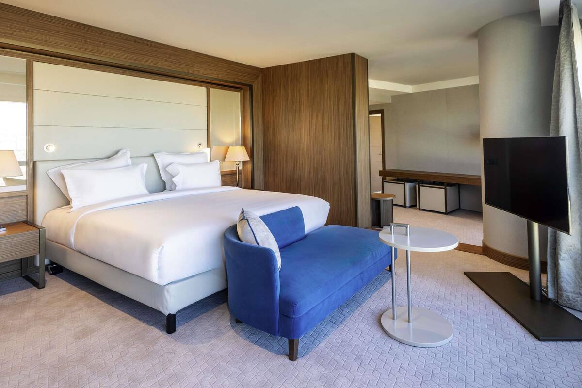 A modern hotel room with a large bed, a blue sofa, a small round table, and a TV.