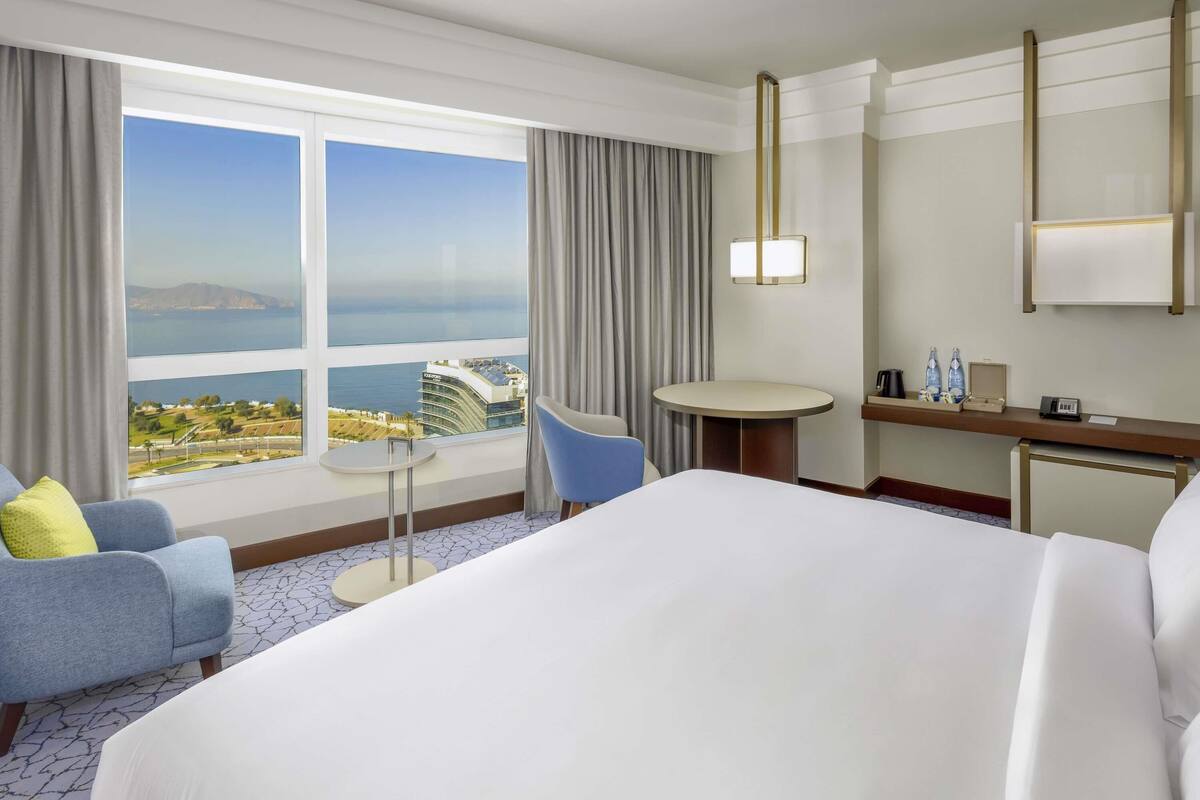 A hotel room with a large bed, a small round table, a chair, a desk with amenities, and a view of the cityscape and water.