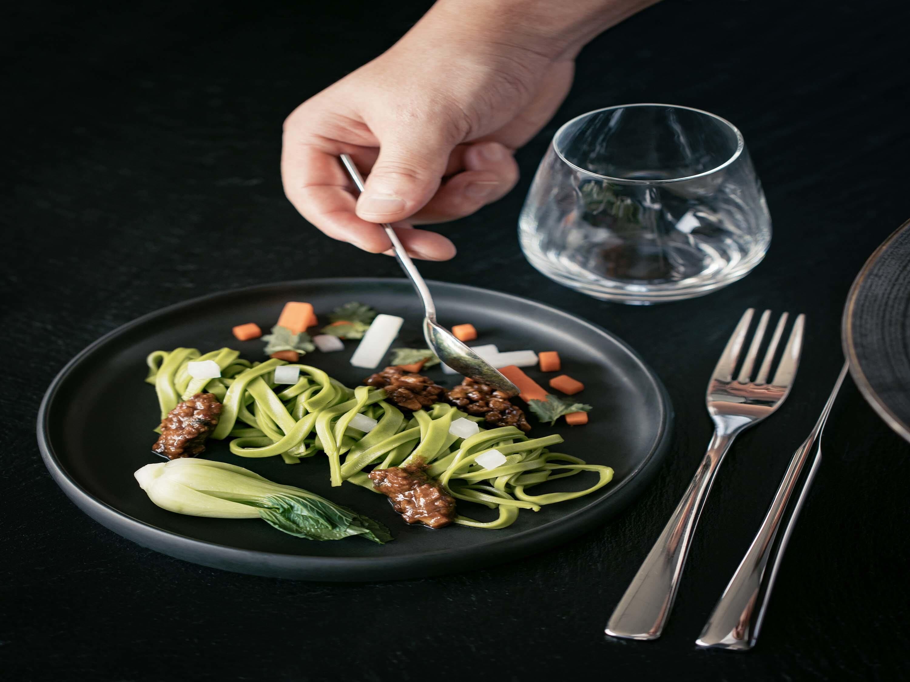 Un plato de fideos con verduras y carne, un vaso de agua y cubiertos sobre una superficie oscura.