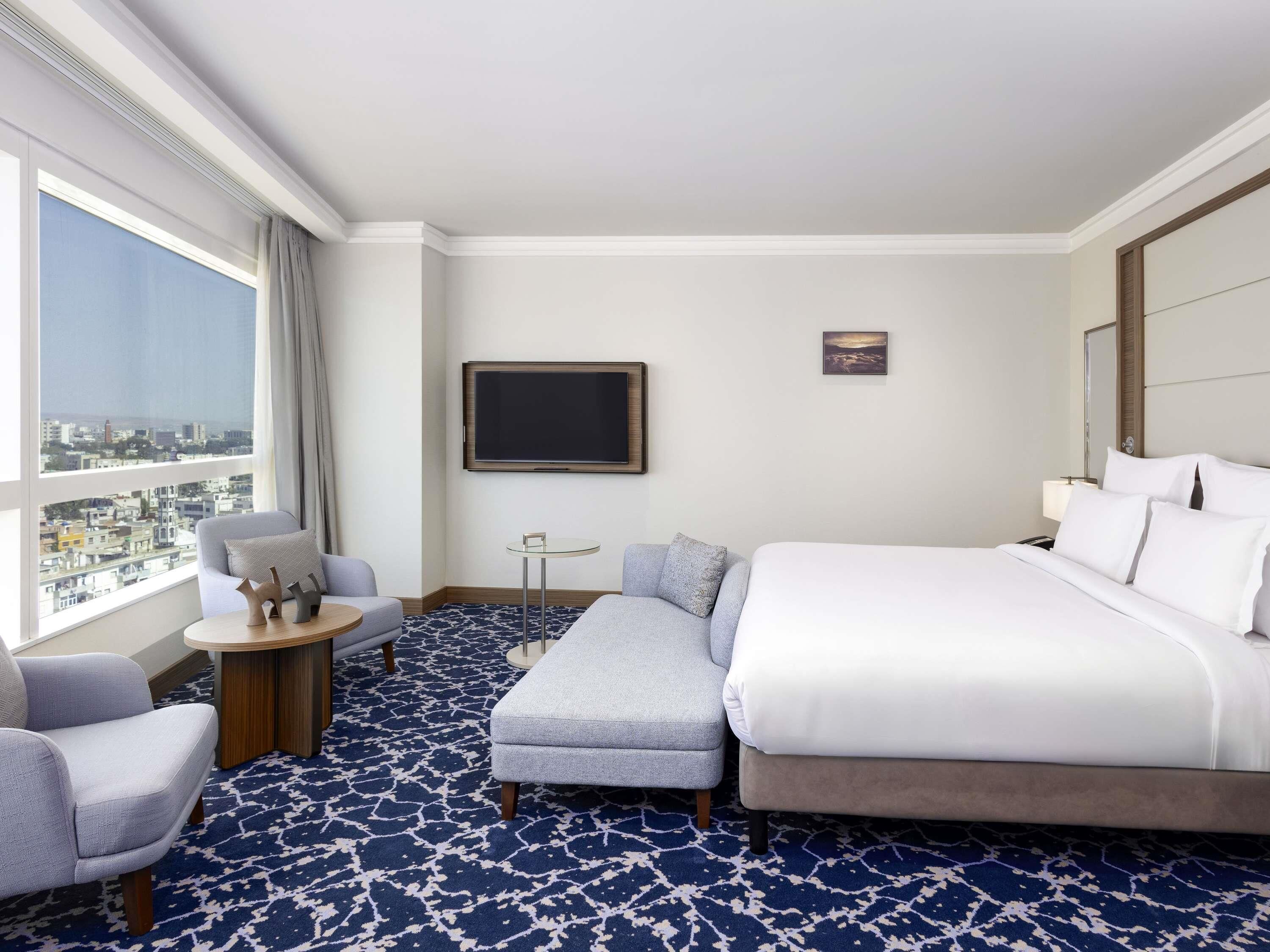 Ein Hotelzimmer mit einem großen Bett, einer Couch, einem kleinen Tisch, einem Fernseher und Blick auf die Stadt.