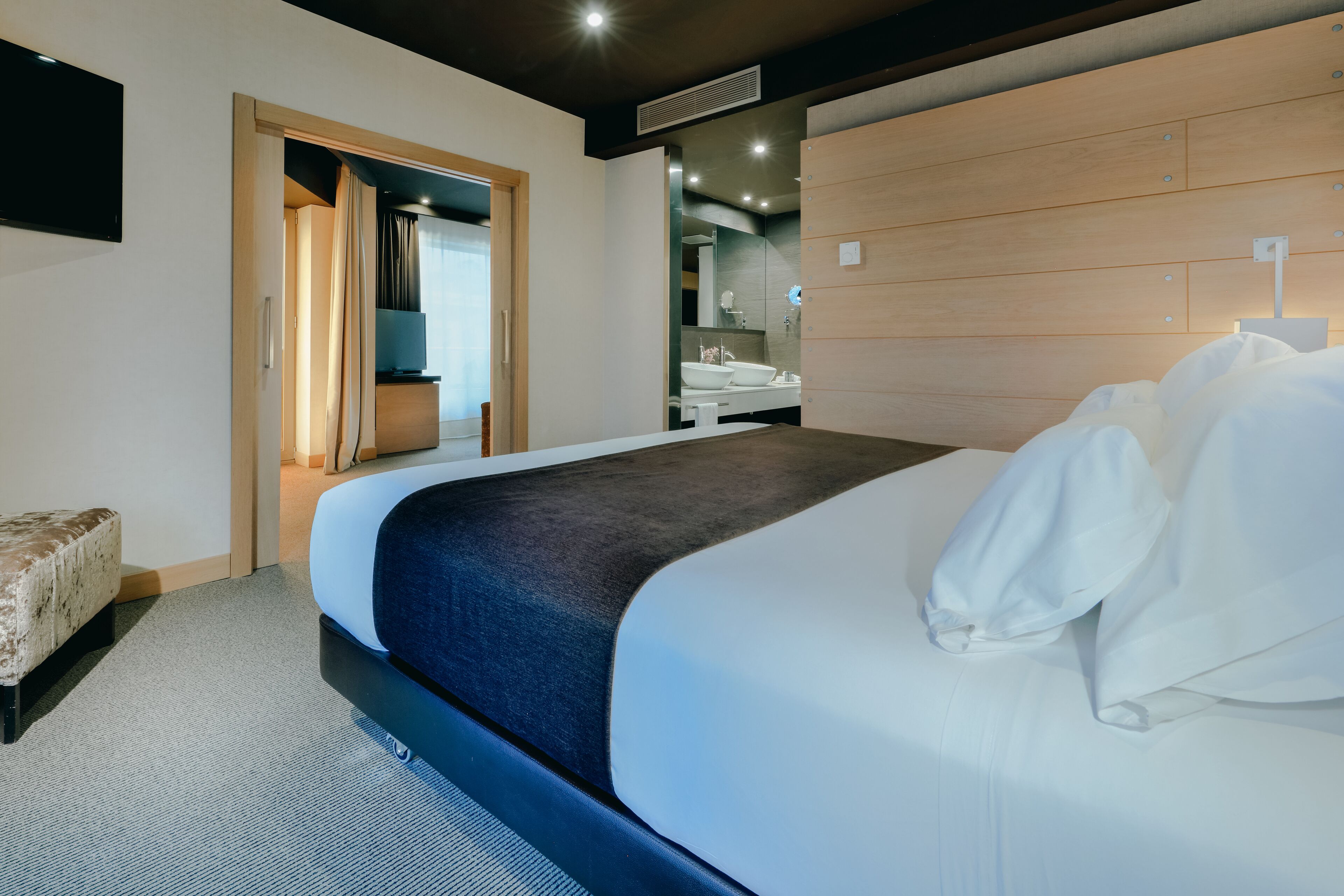 Una habitación de hotel moderna con una cama grande, un cabecero de madera, un baño con espejo y lavabo, y un pequeño espacio de descanso.