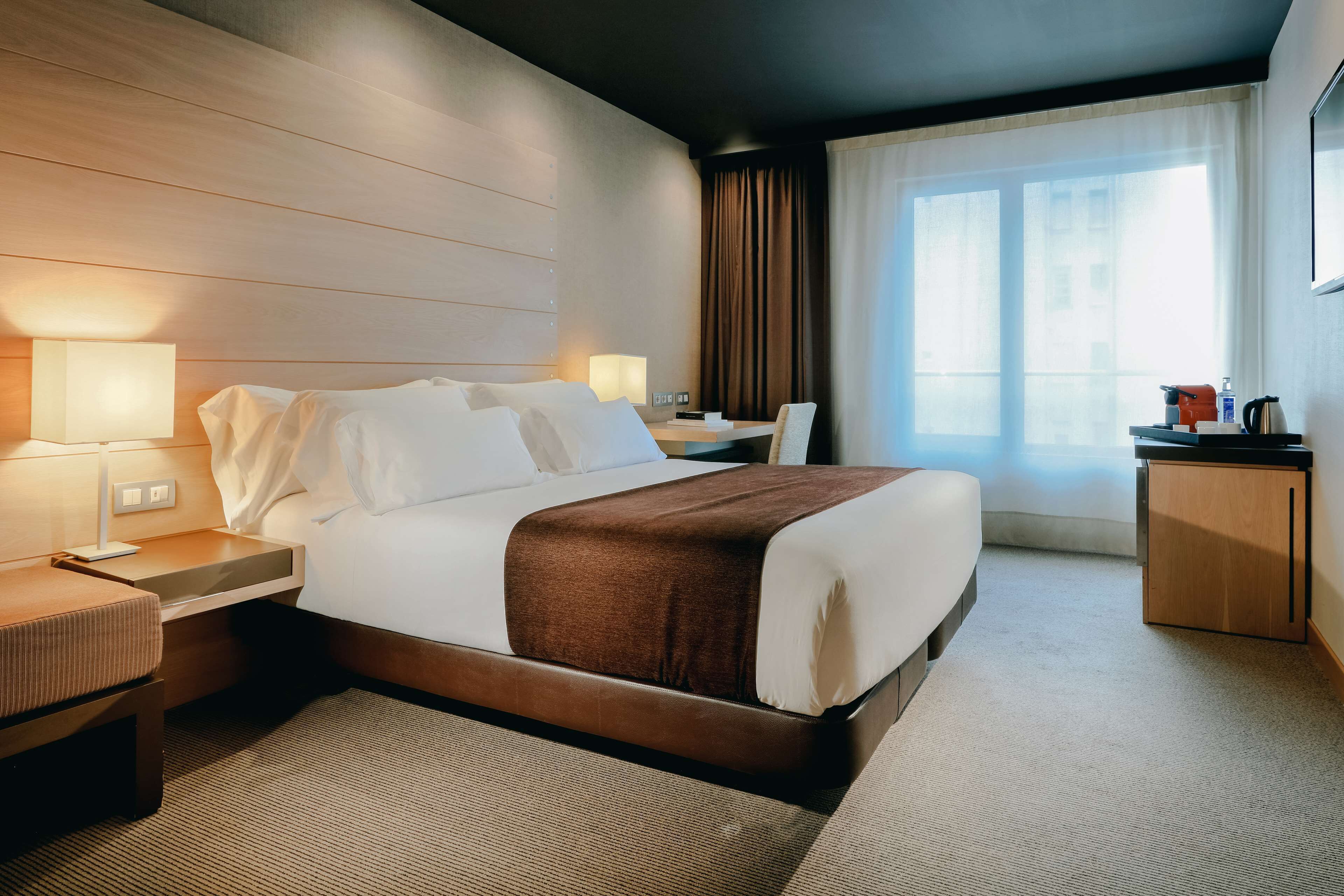 A hotel room with a large bed, bedside tables with lamps, a desk with a chair, a small cabinet, and a large window with curtains.