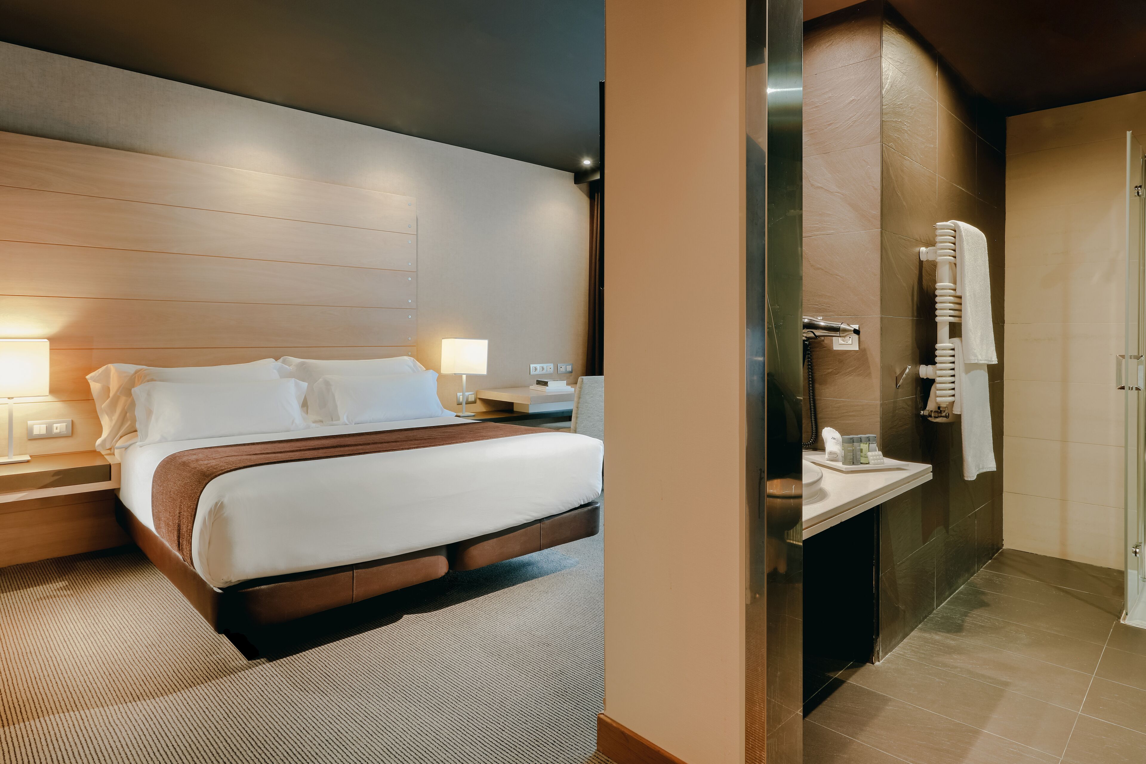 A hotel room with a bed, bedside tables, a nightstand, a lamp, and a bathroom with a sink, mirror, and towel rack.