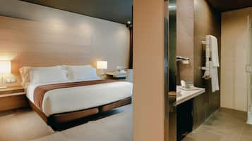 A hotel room with a bed, bedside tables, a nightstand, a lamp, and a bathroom with a sink, mirror, and towel rack.