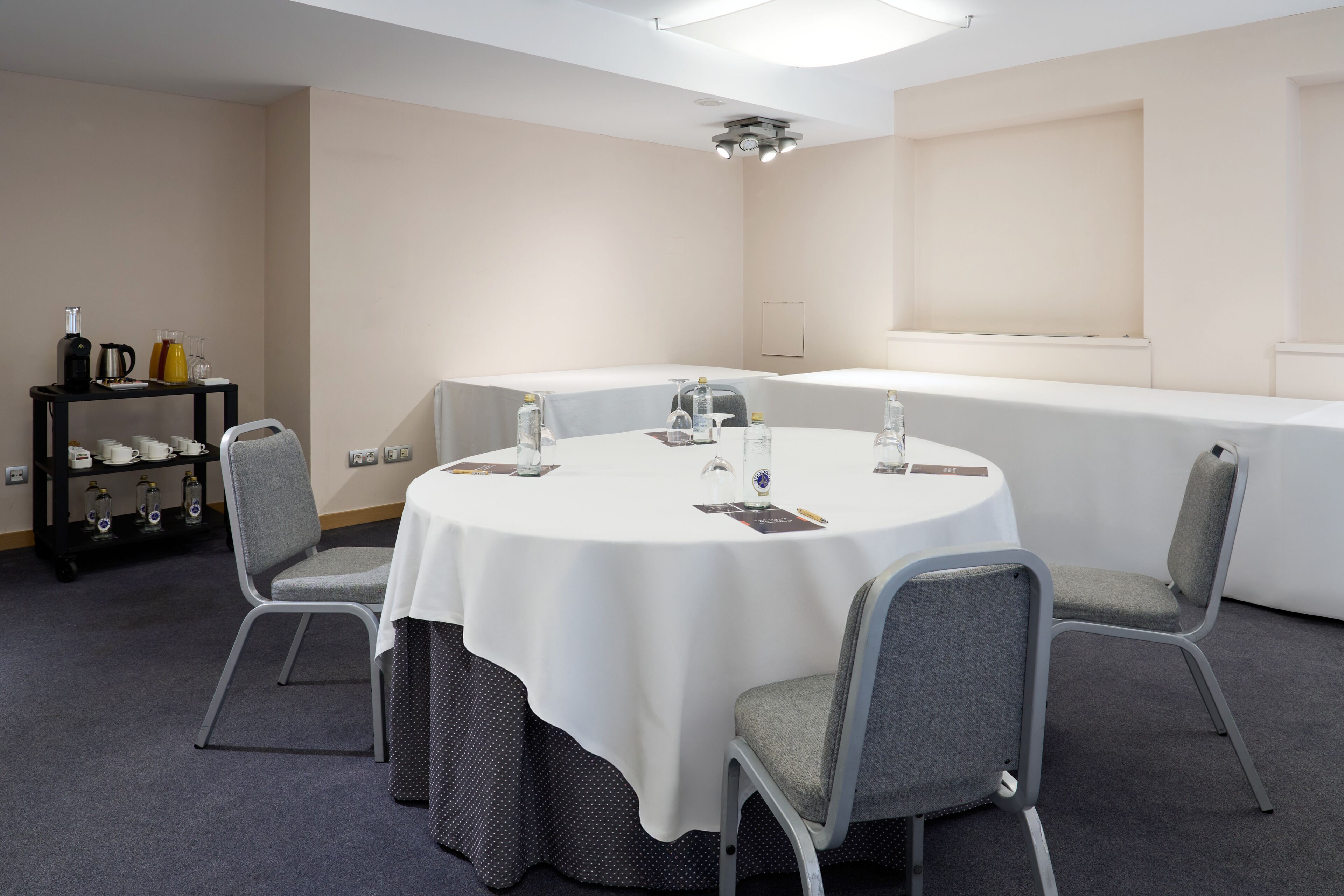 Un salón de conferencias con una mesa redonda, sillas grises, un estante negro con tazas y botellas, y un mantel blanco.