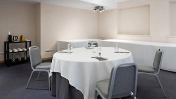 Un salón de conferencias con una mesa redonda, sillas grises, un estante negro con tazas y botellas, y un mantel blanco.
