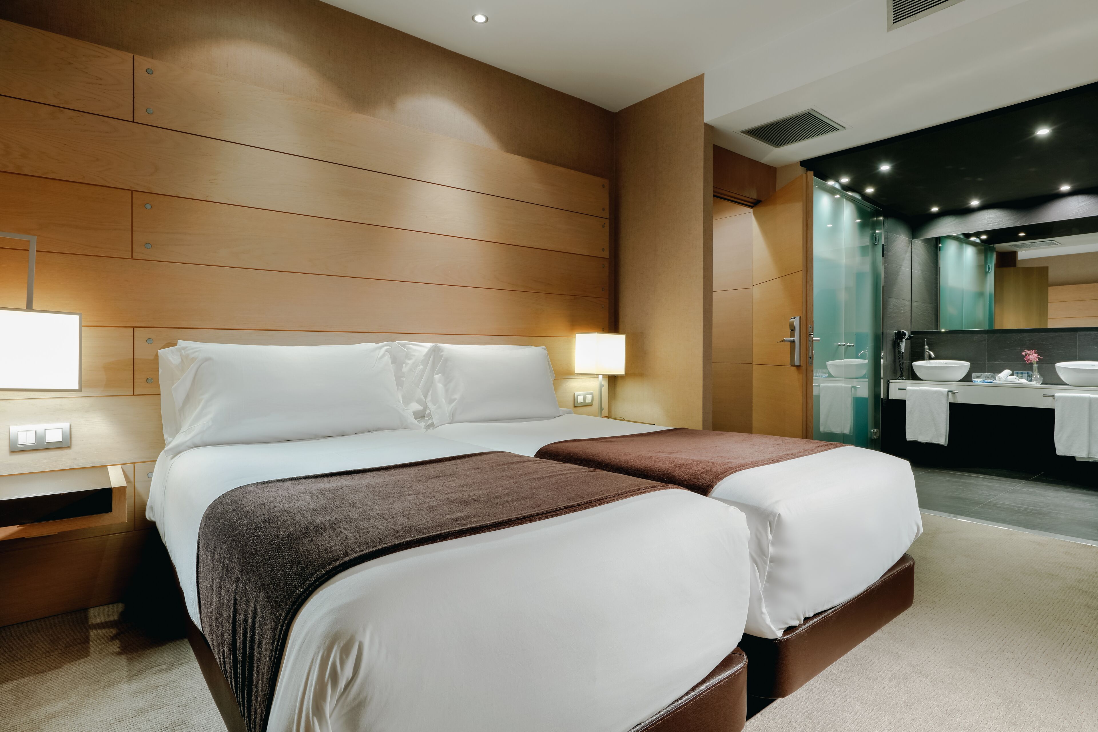 Habitación de hotel con dos camas, cabecera de madera, mesita de noche, escritorio y baño con lavamanos y espejo.
