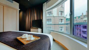 A hotel room with a bed, a TV, a tray with cups, and a view of buildings through a large window.