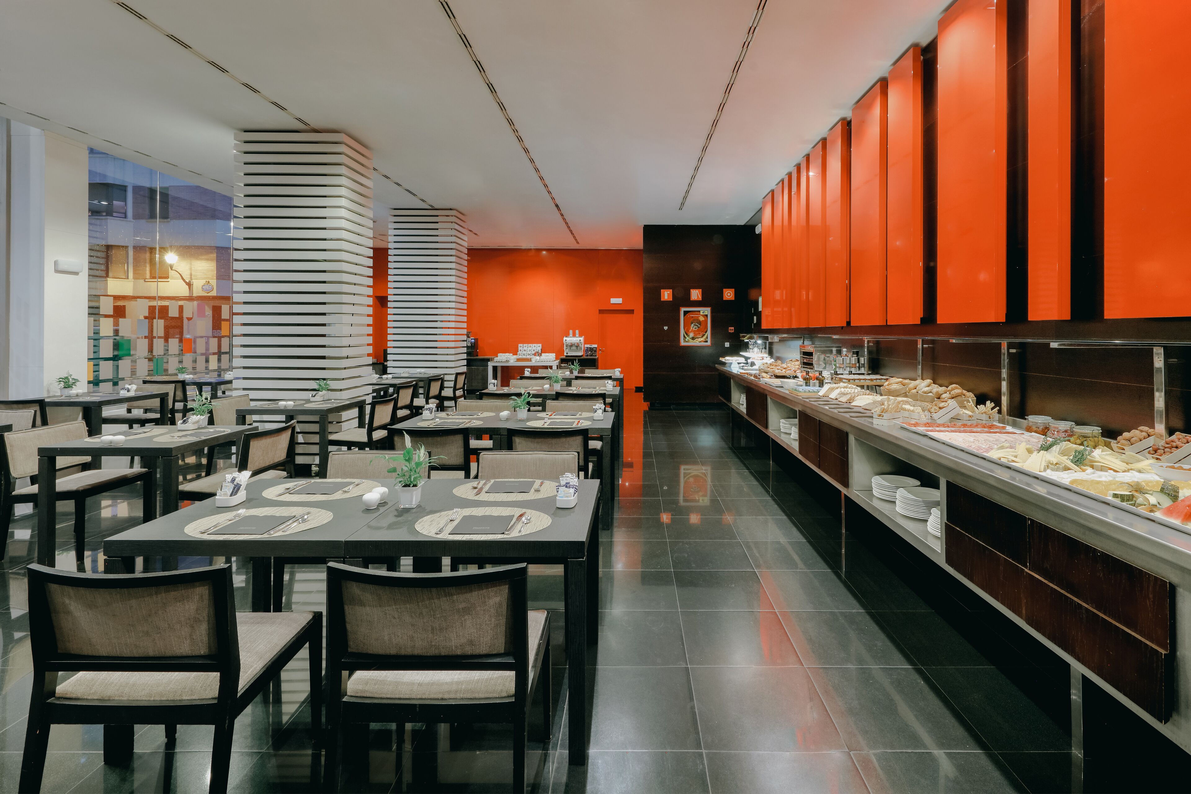 Ein Restaurant im Buffet-Stil mit mehreren Tischen und Stühlen, einer langen Theke mit verschiedenen Speisen und einer auffalligen orangefarbenen Wand.