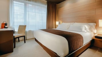 Ett hotellrum med en stor säng, ett skrivbord med en stol, en lampa och ett fönster med gardiner.