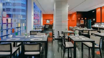 Un restaurante moderno con varias mesas y sillas, un amplio ventanal con vistas a la ciudad y una columna decorativa central.