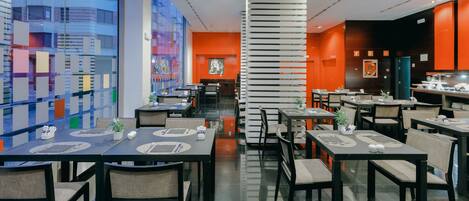 Un restaurante moderno con varias mesas y sillas, un amplio ventanal con vistas a la ciudad y una columna decorativa central.