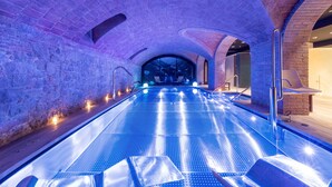 An indoor swimming pool with a stone wall and arched ceiling, featuring a long, narrow pool with a blue floor and a seating area on one side. - Hotel 1898 (Barcelona)