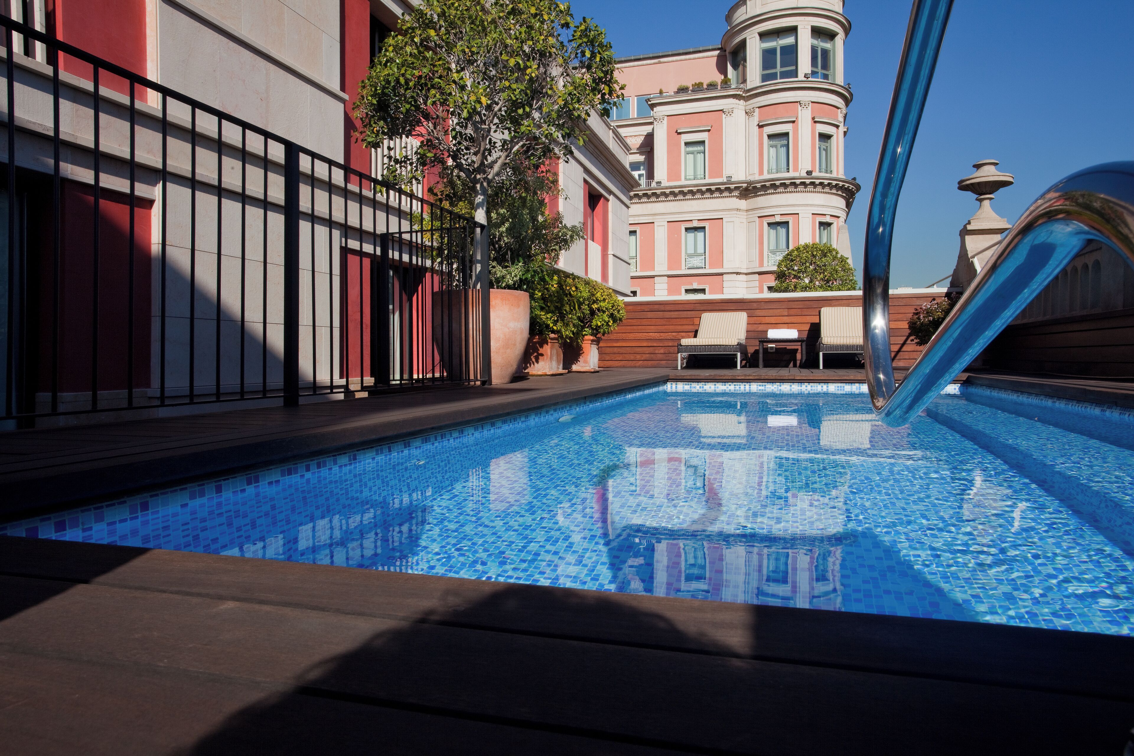 Una piscina con baldosas azules brillantes, rodeada por un muelle de madera y una barandilla de metal. Al fondo, se observa un edificio con fachada rosa y una torre.
