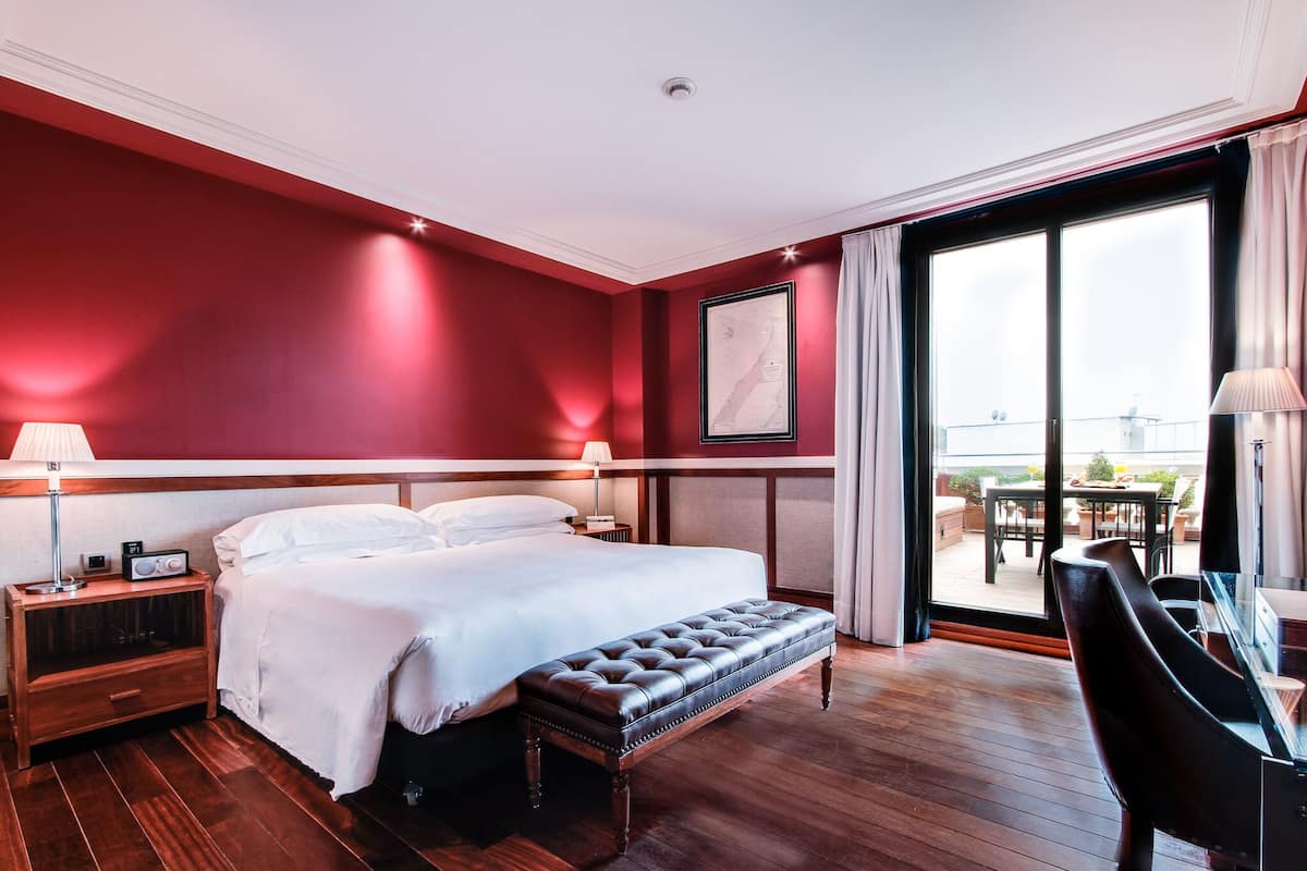 A hotel room with a large bed, a wooden bench, a desk, and a balcony with outdoor furniture.