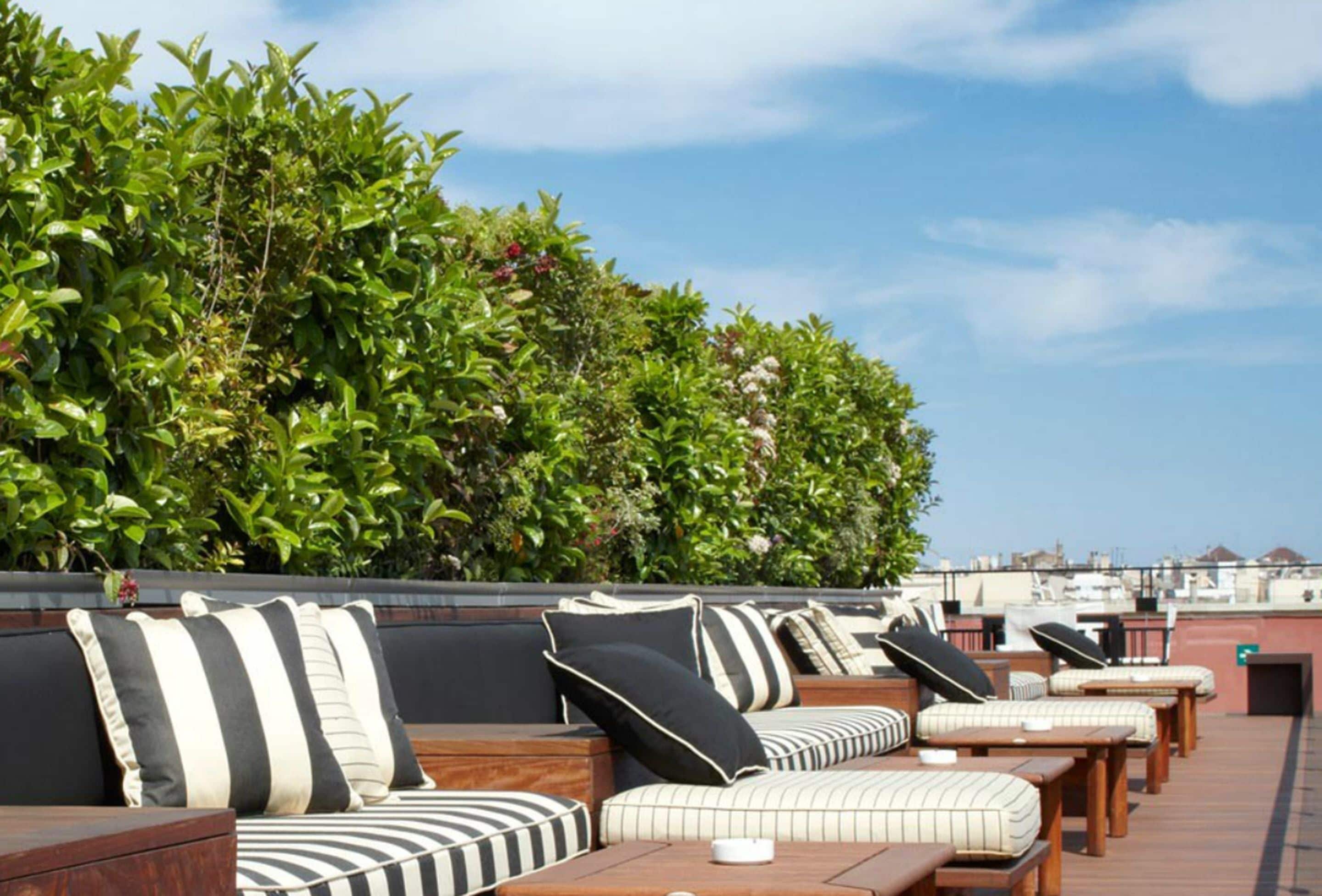 A rooftop lounge area with striped cushions and wooden tables, surrounded by lush greenery.
