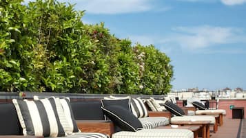 A rooftop lounge area with striped cushions and wooden tables, surrounded by lush greenery.