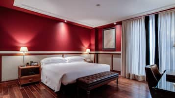 Ein Hotelzimmer mit einem großen Bett, zwei Nachttischen mit Lampen, einer Holzbank, einem Esstisch und einem großen Fenster mit Vorhängen.