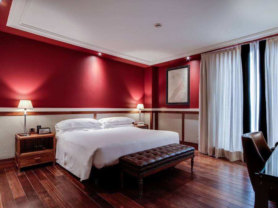 Ein Hotelzimmer mit einem großen Bett, zwei Nachttischen mit Lampen, einer Holzbank, einem Esstisch und einem großen Fenster mit Vorhängen.