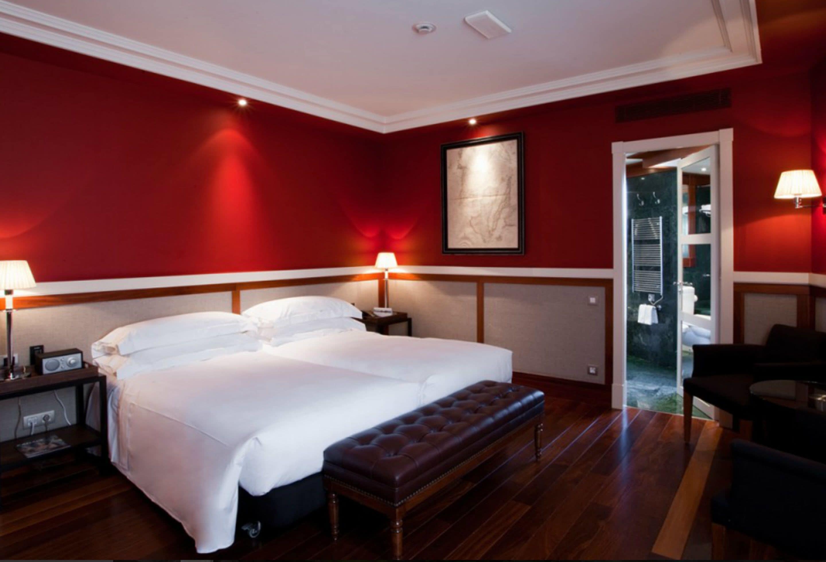A hotel room with a large bed, two bedside lamps, a wooden bench, and a red wall.