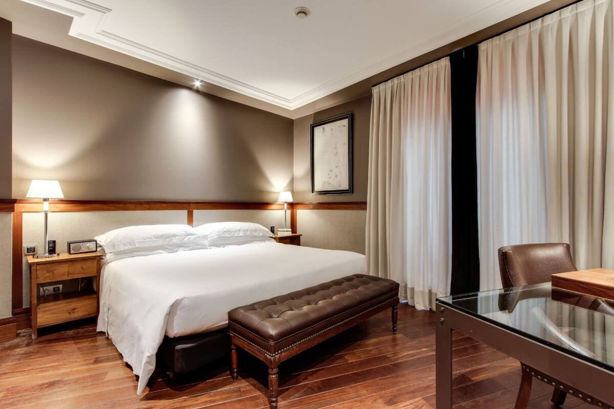 A hotel room with a large bed, a wooden bench, a glass table, and a desk.