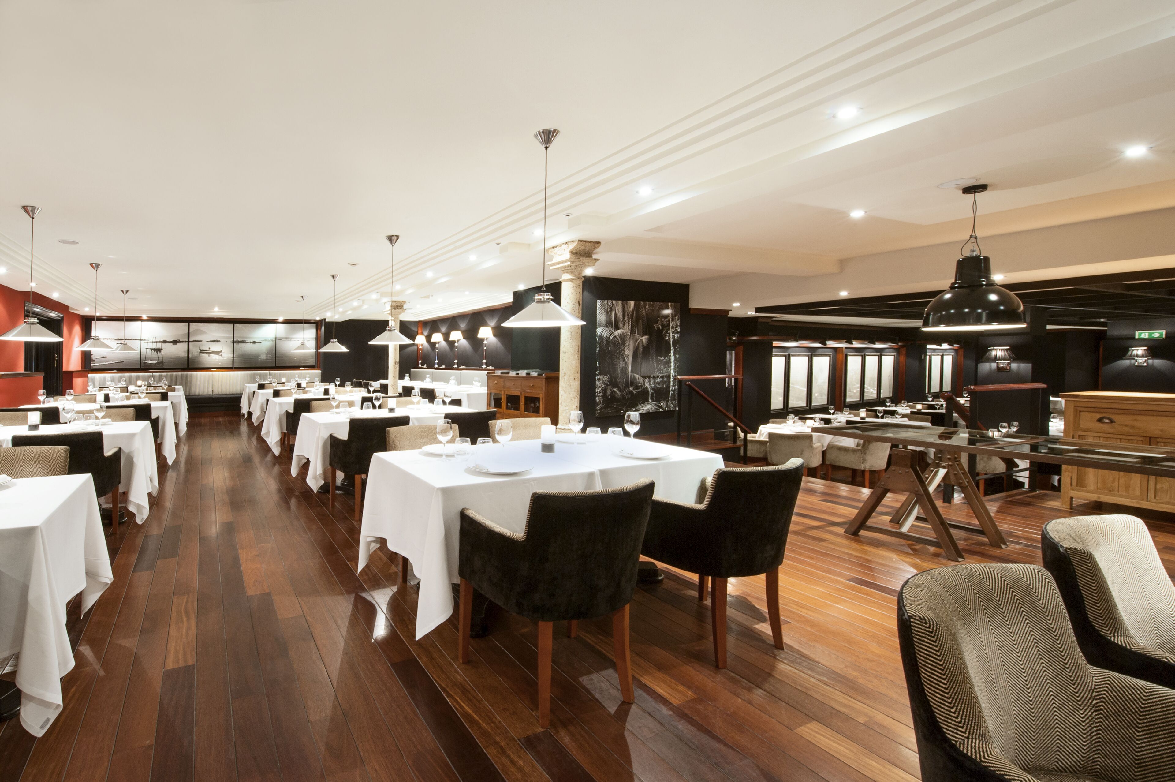 A spacious restaurant with multiple dining tables, modern pendant lighting, and a polished wooden floor.