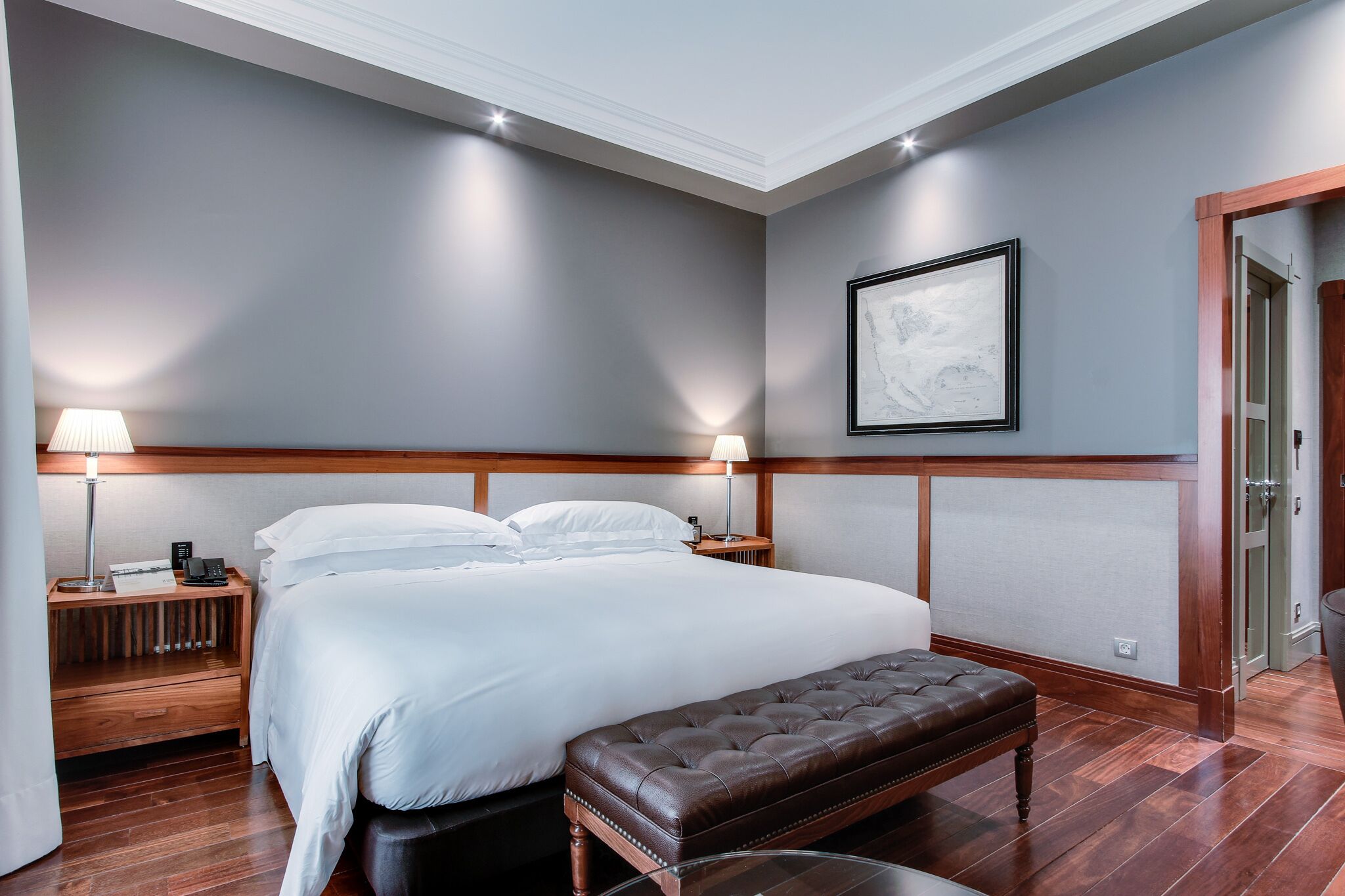 Une chambre d ’hôtel avec un grand lit, deux lampes de chevet, un banc en bois et une œuvre d’art encadrée au mur.
