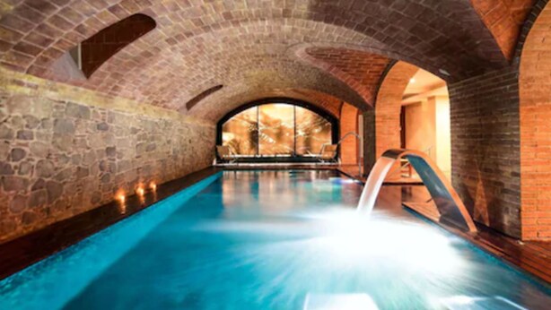 An indoor swimming pool with a brick arch ceiling and a view of a forest through a large window. - Hotel 1898 (Barcelona)