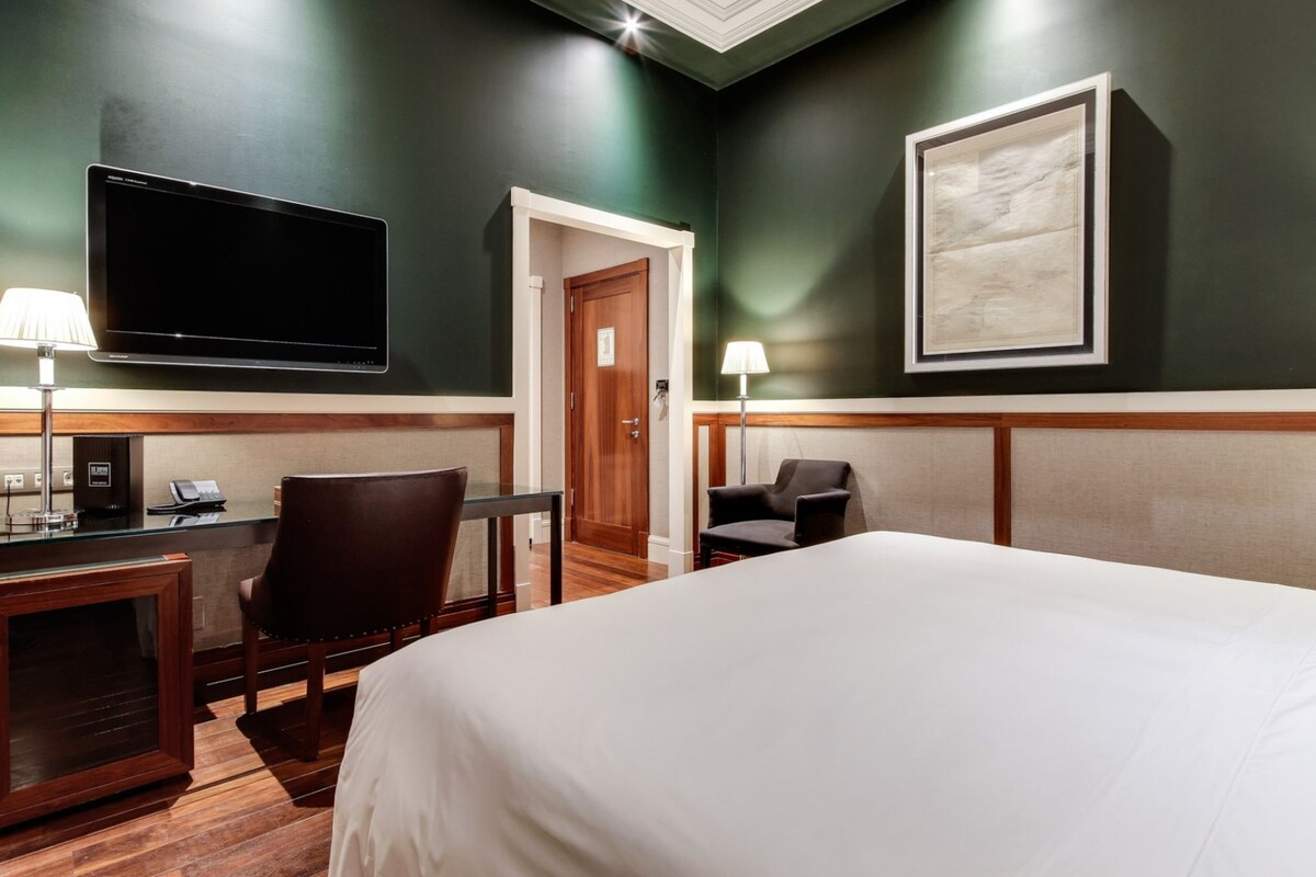 A hotel room with a bed, a desk with a telephone, a chair, a television, and a framed picture on the wall.