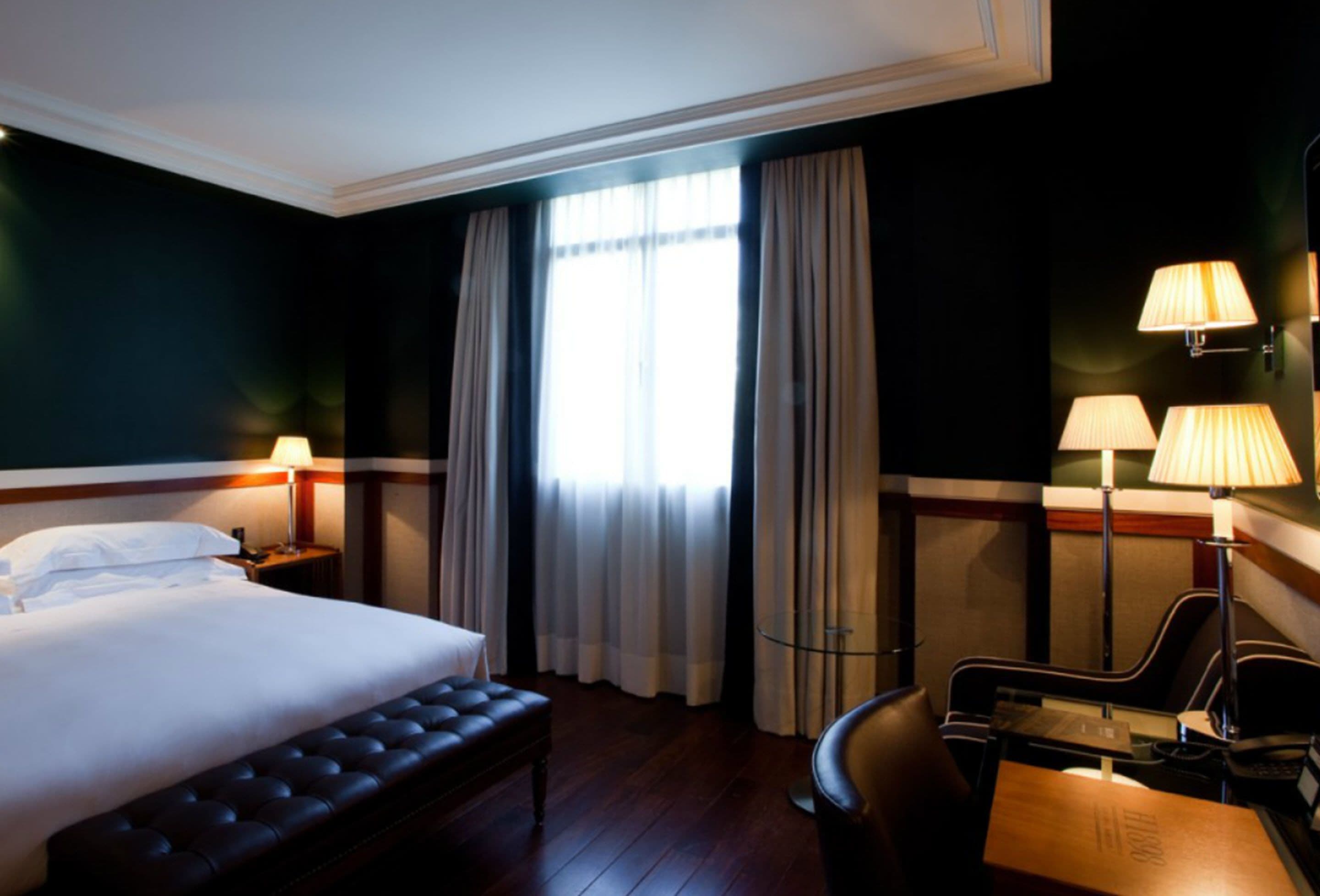 Una habitación de hotel con una cama grande, un banco, dos mesitas de noche con lámparas y un escritorio con silla.
