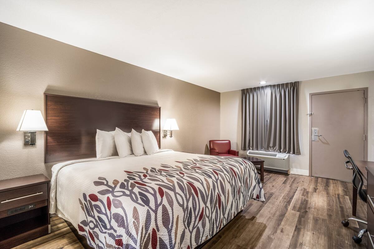 A hotel room with a large bed, a desk, a chair, a window with curtains, and a door.