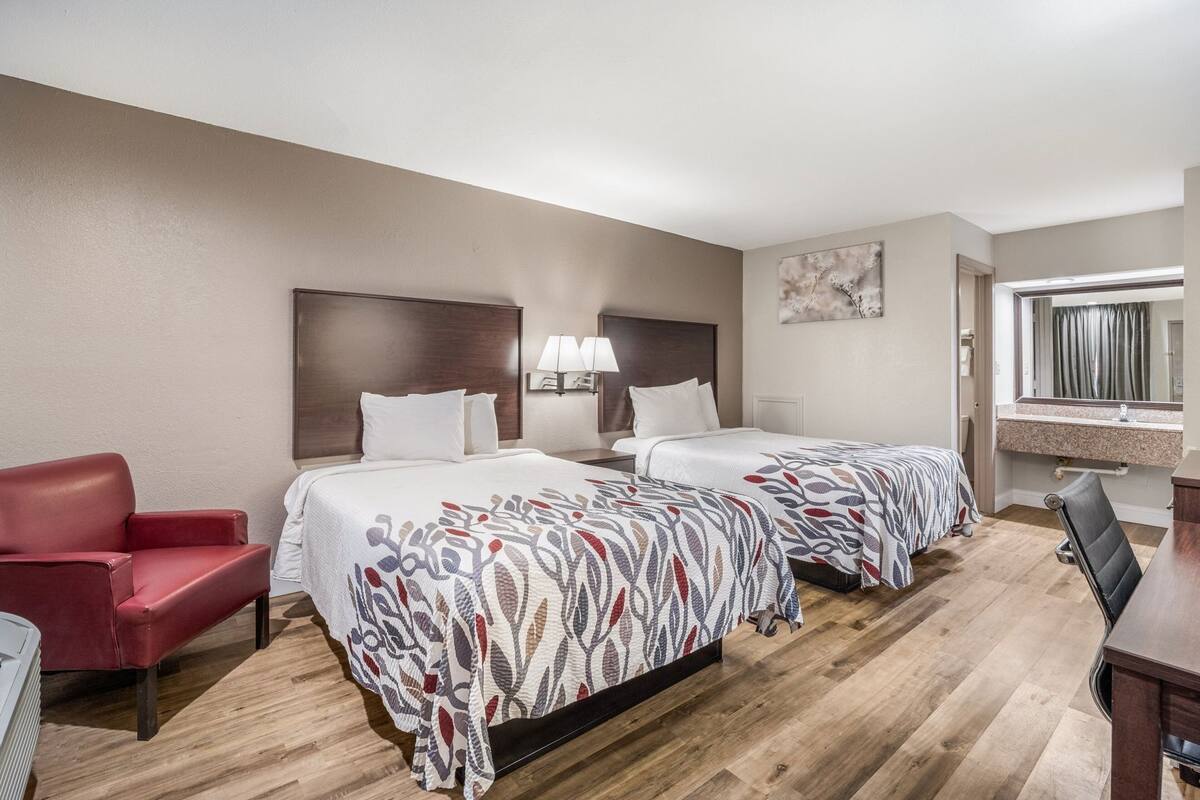 A hotel room with two beds, a red armchair, a desk, and a bathroom with a mirror.