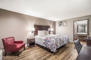 A hotel room with a bed, a red chair, a desk, a mirror, and a wall-mounted lamp. - Red Roof Inn Sumter (Sumter)