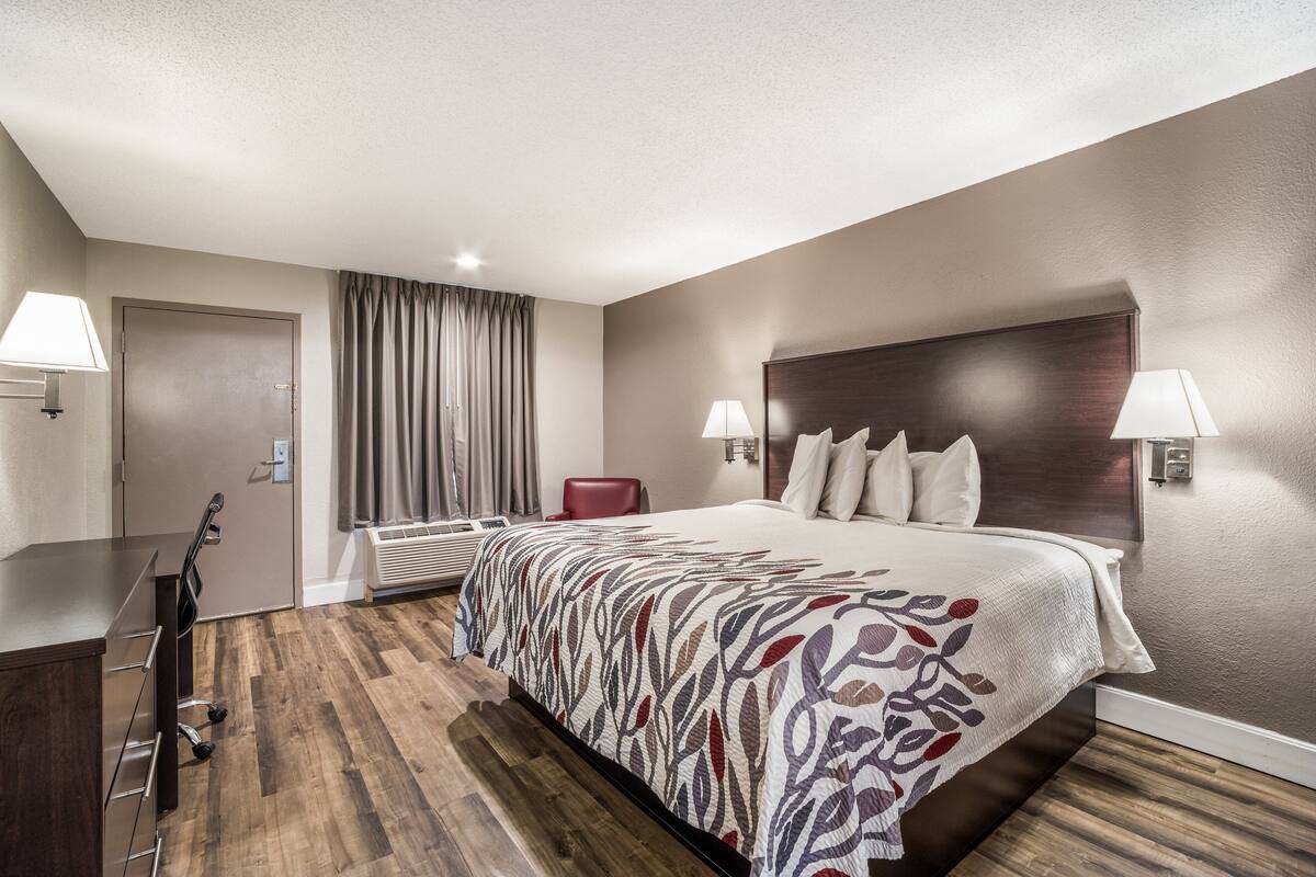 A hotel room with a large bed, two bedside lamps, a wooden floor, a desk with a chair, and a window with curtains.