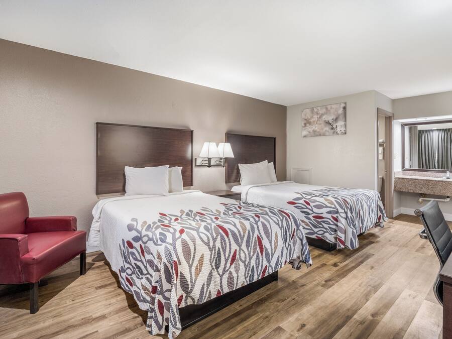 A hotel room with two beds, a red armchair, a desk, and a window with curtains.
