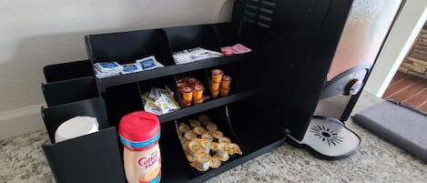 A black storage unit with compartments containing various items such as a bottle of coffee creamer, packaged food items, and a small trash bin.