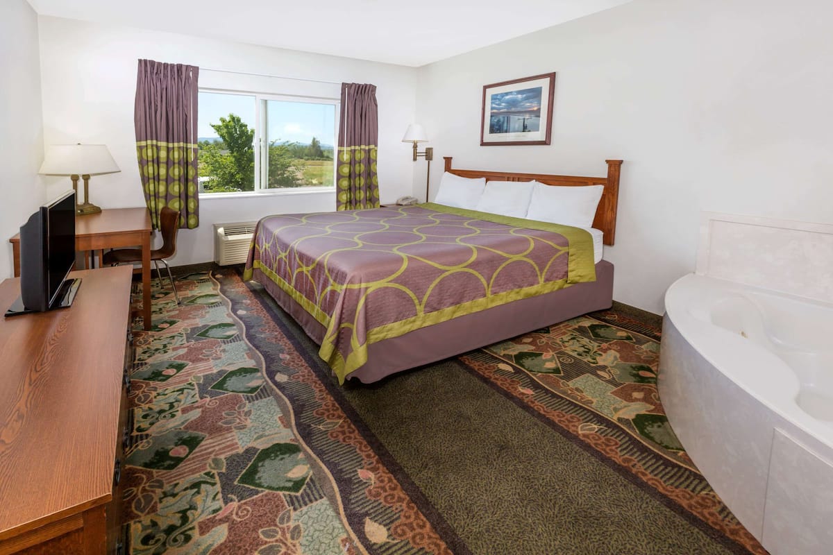 A hotel room with a bed, a desk with a computer, a chair, a window with curtains, a wall-mounted lamp, a framed picture, and a bathtub.