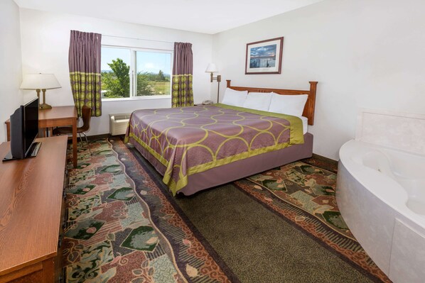 A hotel room with a bed, a desk with a computer, a chair, a window with curtains, a wall-mounted lamp, a framed picture, and a bathtub.