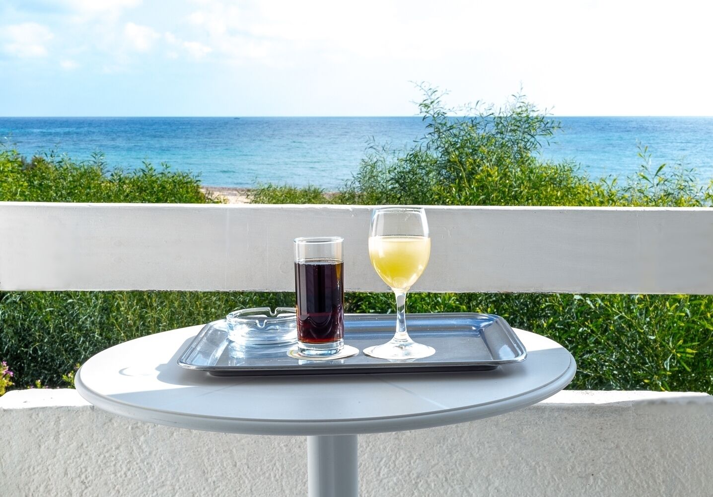 Een tafel met een dienblad waarop een glas donkere vloeistof en een glas witte vloeistof staan, voor een achtergrond van een balkon met uitzicht op zee.