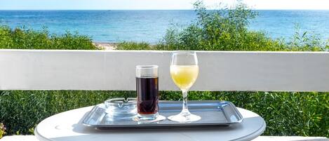 Een tafel met een dienblad waarop een glas donkere vloeistof en een glas witte vloeistof staan, voor een achtergrond van een balkon met uitzicht op zee.